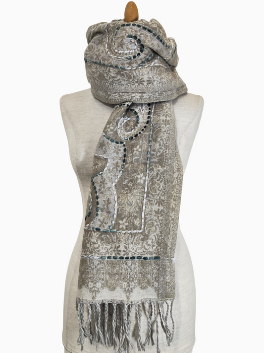 Ein luxuriöser Wendeschal aus beiger Lammwolle, verziert mit detaillierter weißer Paisley-Stickerei und funkelnden grünen Perlenakzenten. Das Design hat weiche Fransen an den Rändern, die ihm einen Hauch von Eleganz verleihen. Der Schal ist sowohl für die Freizeit als auch für formelle Anlässe geeignet und wird auf einer Schaufensterpuppe präsentiert, um die aufwendige Verarbeitung und den leichten Faltenwurf zu betonen. Perfekt zum Überziehen oder als auffälliges Accessoire.