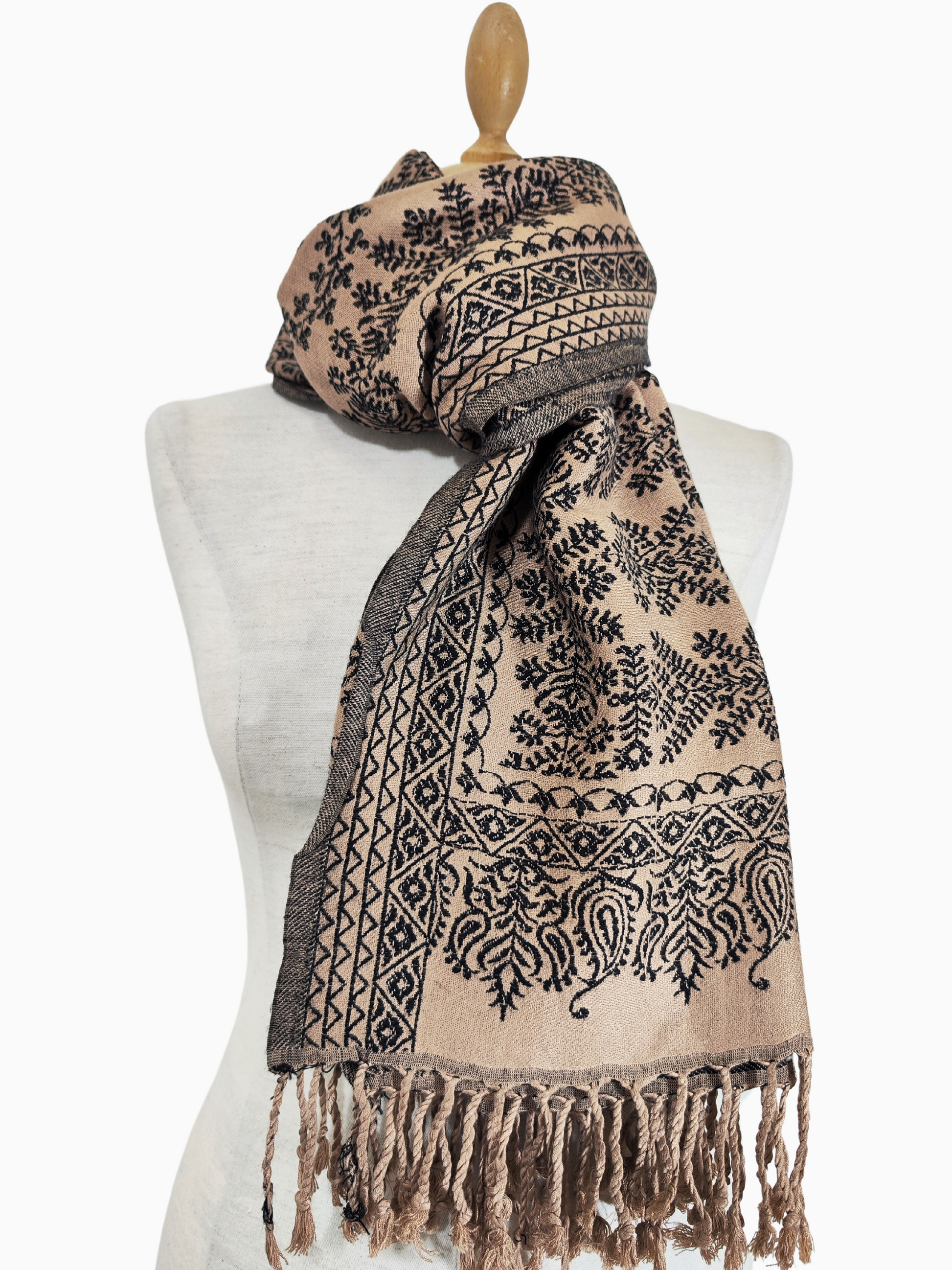 A beige shawl with intricate black floral patterns and fringed edges. A luxurious cotton-cashmere blend shawl in beige with a beautiful pattern of black floral and geometric motifs. The shawl has fringed edges.